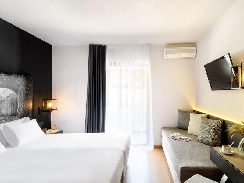 Moderní hotelový pokoj s manželskou postelí, pohovkou, televizí na stěně a světlým balkonovým oknem.