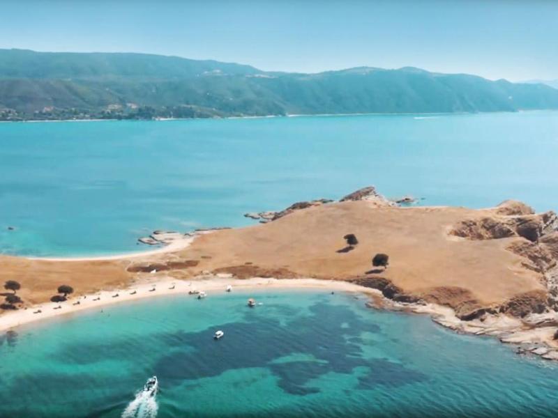 Letecký pohled na skalnatý poloostrov s čistou modrou vodou a horami v pozadí.