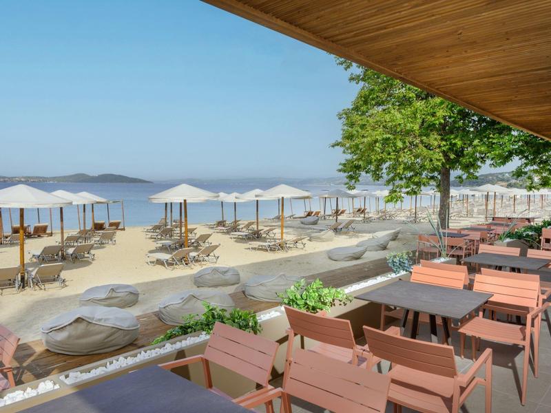 Výhled na pláž z kryté terasy s stoly, židlemi, slunečníky a písečnou pláží.