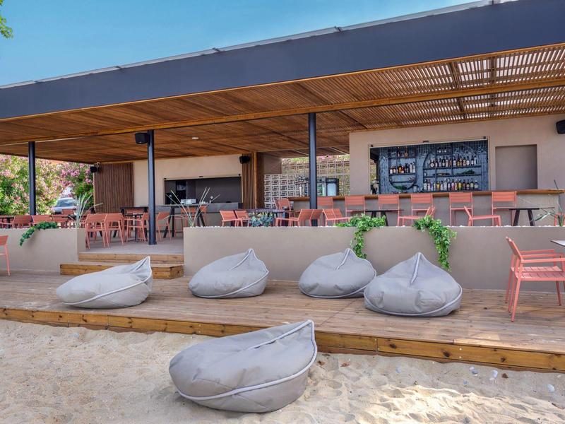 Plážový bar s dřevěnou střechou, sedacími vaky a stolky na písčité pláži za slunečného počasí.