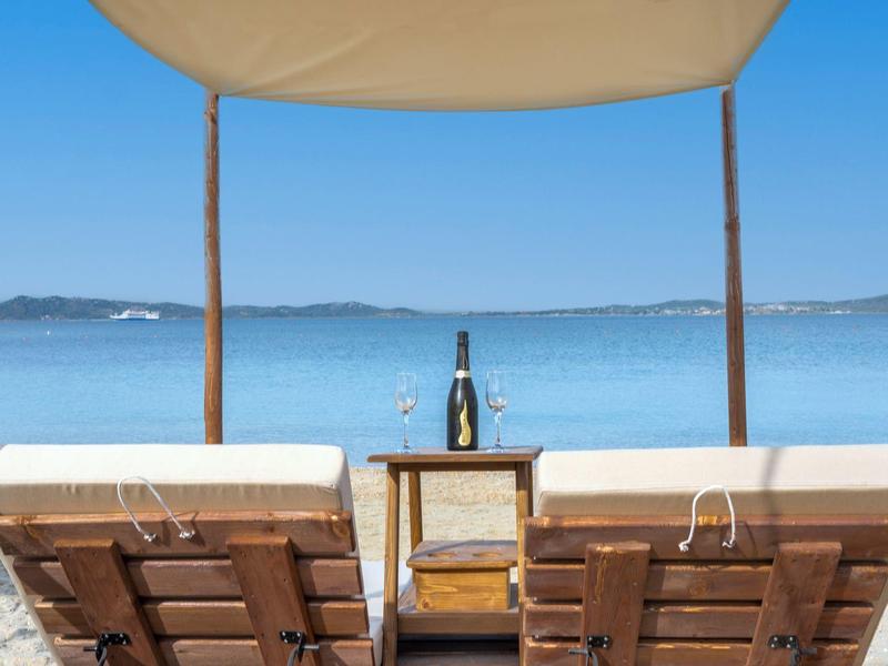 Dvě lehátka pod přístřeškem s výhledem na klidné moře a vínem na malém stolku