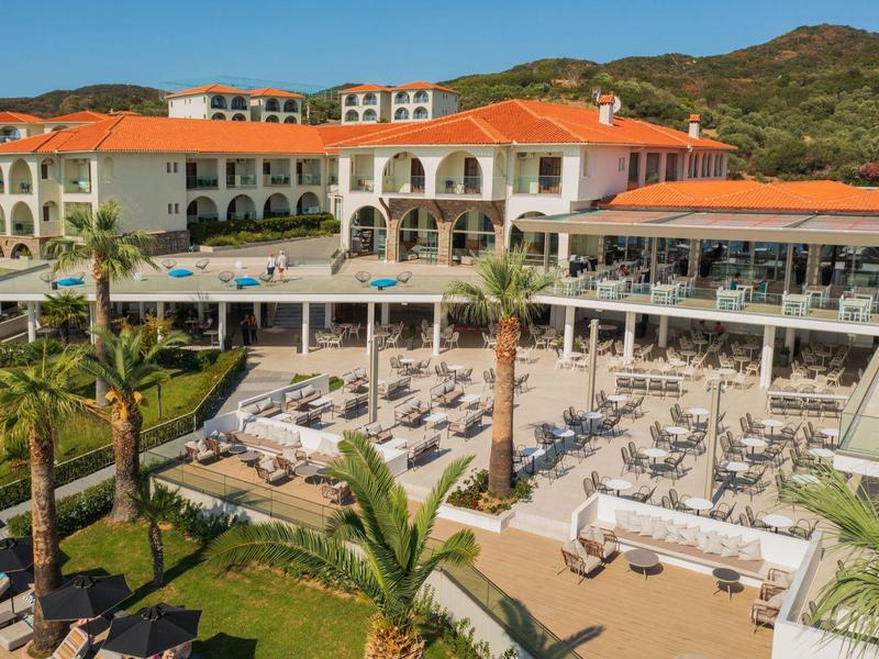 Velký hotel s bazénem, sluneční terasou a palmami před kopcovitou krajinou za jasné oblohy.