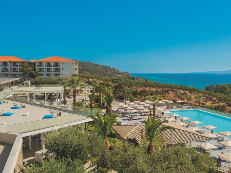 Pohled na resort s bazénem, slunečníky a mořem v pozadí za jasné oblohy