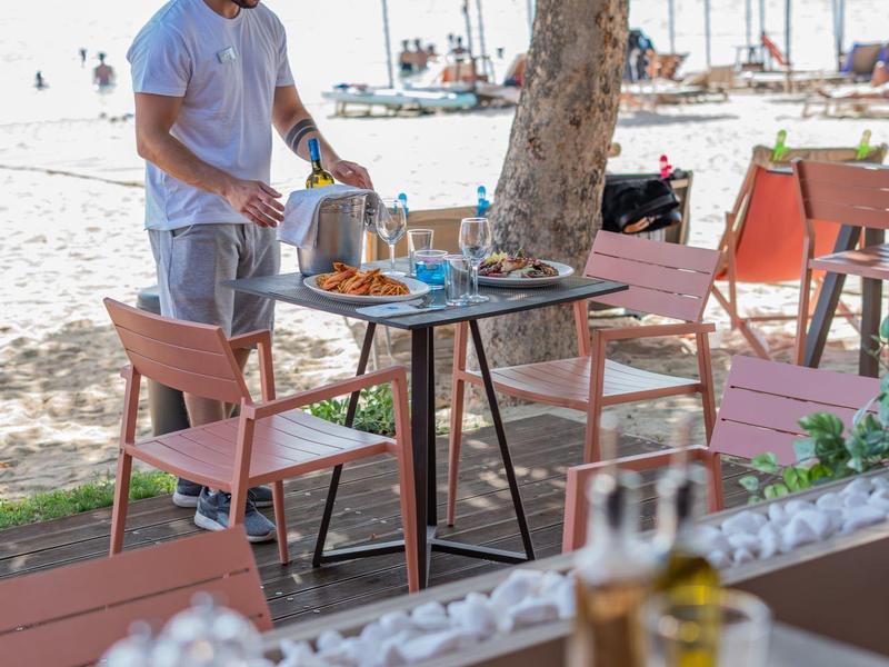 Muž stojí u venkovního stolu na pláži pod stromem s židlemi a slunečníky v pozadí.