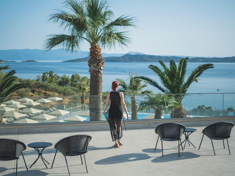 Žena stojí na terase s výhledem na moře a palmy.