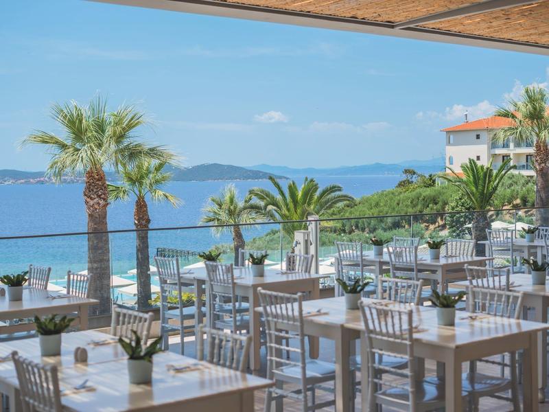 Terasa restaurace s stoly a židlemi, palmami a výhledem na moře a hory.