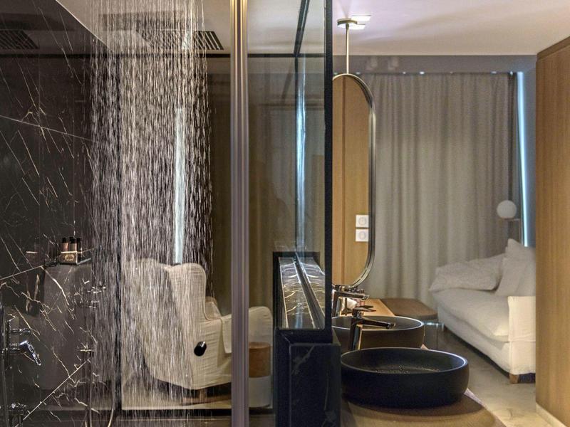 Moderní hotelový pokoj se skleněnou sprchou, pohodlným křeslem a bílou postelí.