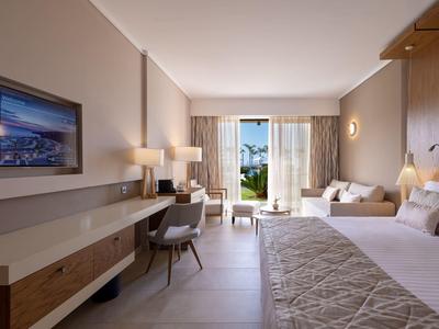 Světý hotelový pokoj se dvěma postelemi, psacím stolem a otevřenými dveřmi na balkon s výhledem na moře.