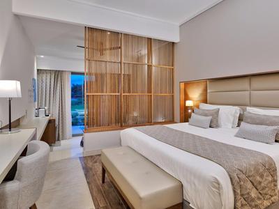 Moderní hotelový pokoj s velkou postelí, béžovými tóny, dřevěnou příčkou a posezením.