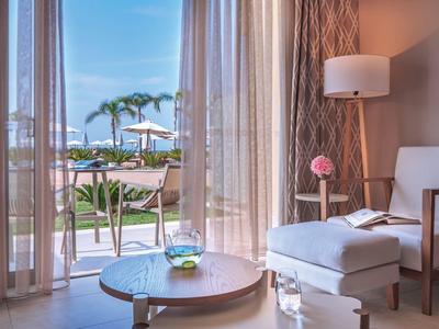 Pohodlný hotelový pokoj s křeslem, konferenčním stolkem a skleněnými dveřmi na slunnou terasu u bazénu s palmami.