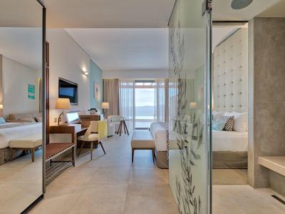 Moderní hotelový pokoj s postelemi, posezením, skleněnou sprchou a balkonem s výhledem na oceán.