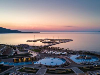 Pohled na západ slunce nad pobřežním letoviskem s bazény, marinou a vzdálenými kopci u klidného moře.