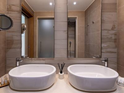 Moderní koupelna s dvojitými umyvadly, béžovými dlaždicemi a matnými skleněnými dveřmi sprchy.