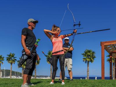 Tres personas practican tiro con arco en un campo de césped bajo un cielo soleado con palmeras al fondo.