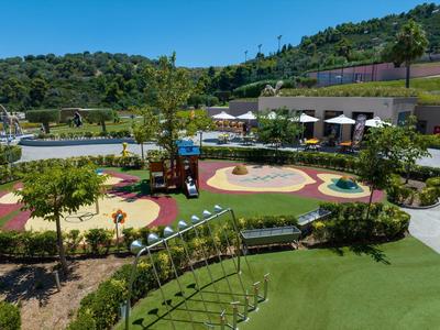 Venkovní hřiště s skluzavkami, houpačkami a slunečníky v zelené hotelové zahradě na kopci.