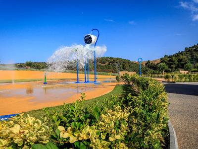 Modré vodní fontány rozprašující mlhu na slunném parku s zelenými keři a jasnou oblohou.