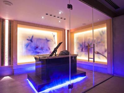 Moderní spa místnost s kamenným stolem a fialovým ambientním osvětlením za skleněnými stěnami.