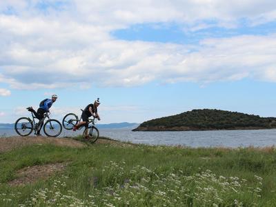 Dos ciclistas de montaña en una colina cubierta de hierba con vista a un lago bajo un cielo nublado.