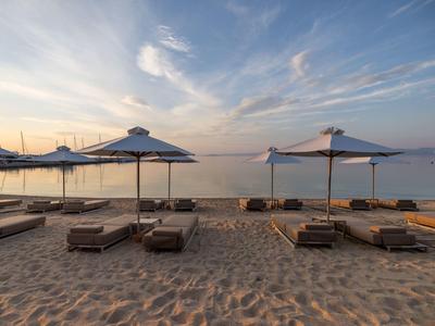 Pláž s lehátky a slunečníky směřující k klidnému moři pod částečně oblačnou oblohou při západu slunce.