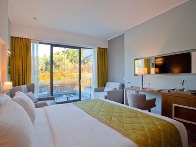 Moderní hotelový pokoj s velkou postelí, zelenou přikrývkou, posezením a velkým oknem s výhledem na zeleň.