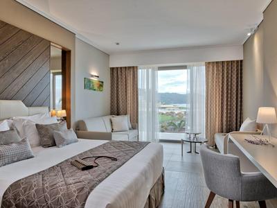 Světý hotelový pokoj s postelí, pohovkou, psacím stolem a balkonem s výhledem.