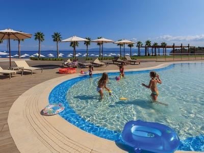 Děti si hrají v čistém venkovním bazénu s lehátky a slunečníky v blízkosti pod modrou oblohou.