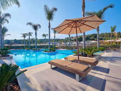 Area piscina del resort soleggiata con lettini, ombrelloni, palme e cielo azzurro.