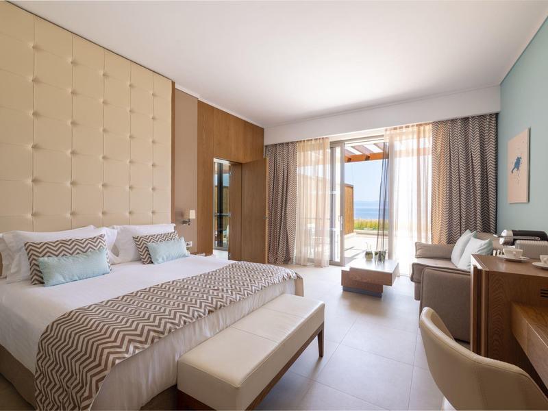 Moderní hotelový pokoj s velkou postelí, béžovým vybavením, psacím stolem a balkonem s výhledem na moře.