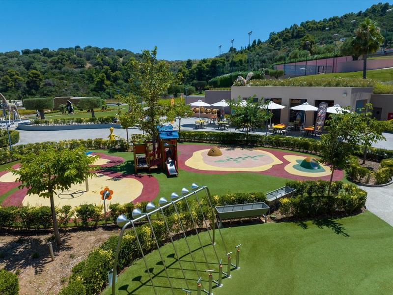 Venkovní hřiště s skluzavkami, houpačkami a slunečníky v zelené hotelové zahradě na kopci.