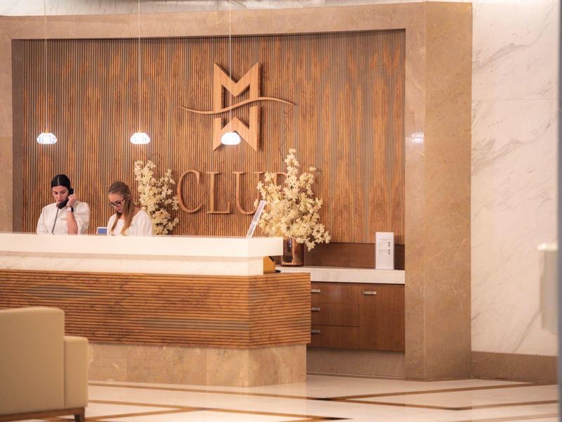 Hotelová recepce s dřevěným pultem, mramorovými stěnami a personálem pomáhajícím hostům.