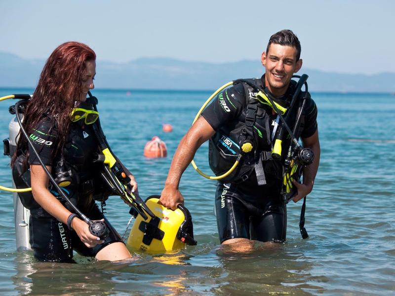 Dva potápěči v oblecích stojí v mělké vodě a drží potápěčské přilby.