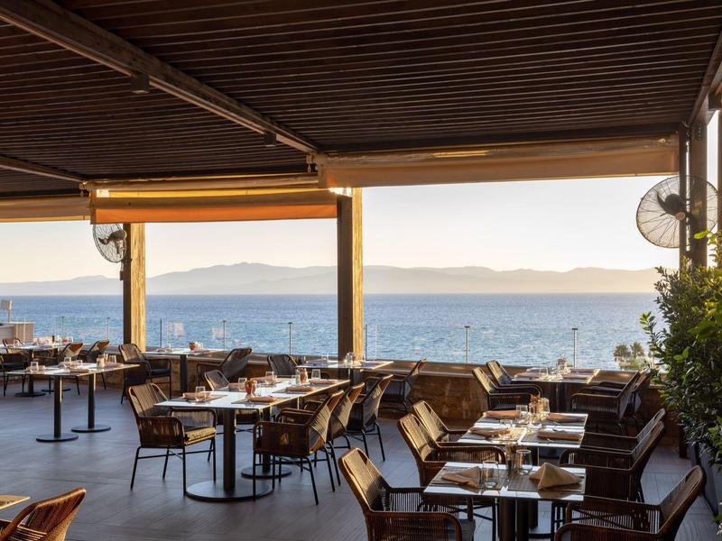 Venkovní jídelní prostor s výhledem na moře, dřevěnou střechou a prostřenými stoly a židlemi.