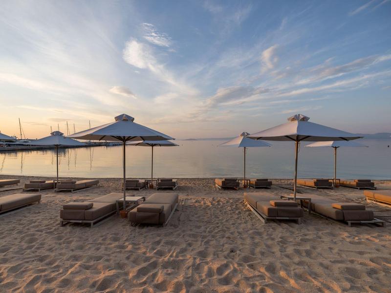 Pláž s lehátky a slunečníky směřující k klidnému moři pod částečně oblačnou oblohou při západu slunce.