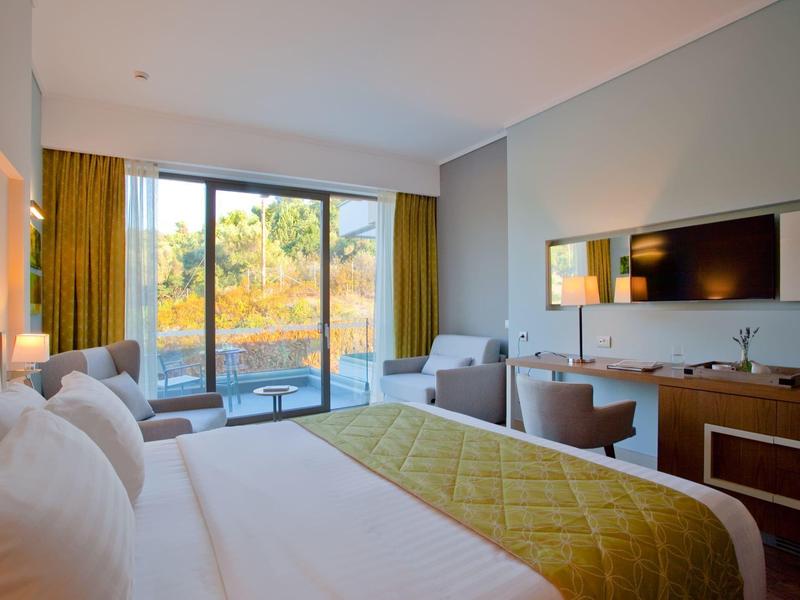 Moderní hotelový pokoj s velkou postelí, zelenou přikrývkou, posezením a velkým oknem s výhledem na zeleň.