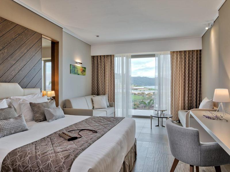 Světý hotelový pokoj s postelí, pohovkou, psacím stolem a balkonem s výhledem.