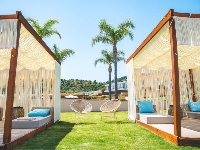 Venkovní relaxační prostor s dřevěnými altánky, závěsnými židlemi a palmami za slunečného dne.