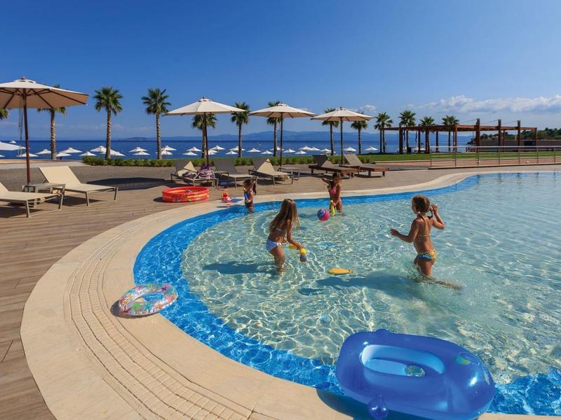 Děti si hrají v čistém venkovním bazénu s lehátky a slunečníky v blízkosti pod modrou oblohou.