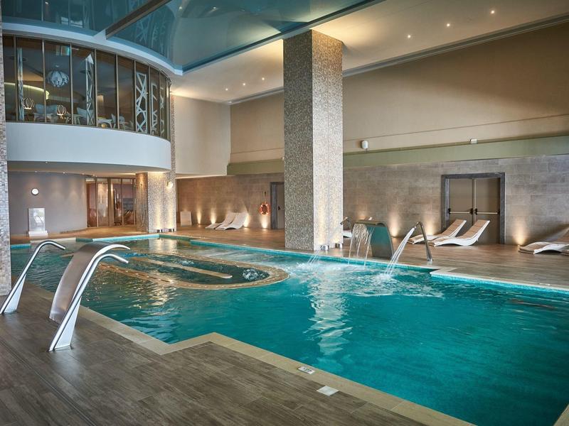 Vnitřní bazén s lehátky, kamennými sloupy a zakřiveným balkonem nad spa.