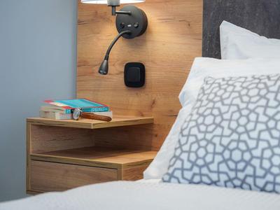 Holz-Nachttisch mit Büchern, Leselampe und modernem Wandschalter neben einem Bett mit grauem Kissen.