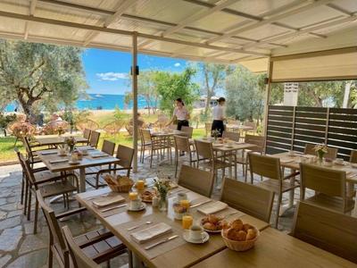 Offener Restaurantbereich mit Holztischen und Blick auf das Meer und Olivenbäume.