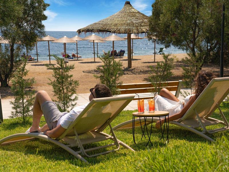 Dwie osoby odpoczywają na leżakach na trawie z widokiem na plażę i morze.