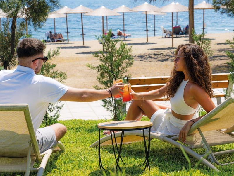 Para siedzi na leżakach na trawie, stukając się napojami przy plaży z widokiem na morze.