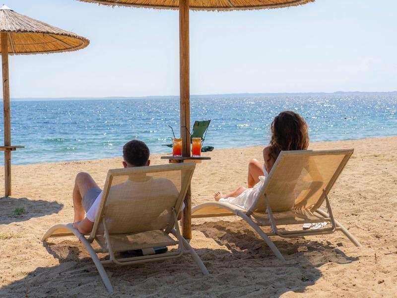 Dwie osoby odpoczywają na leżakach pod parasolami ze słomy na piaszczystej plaży z widokiem na morze.