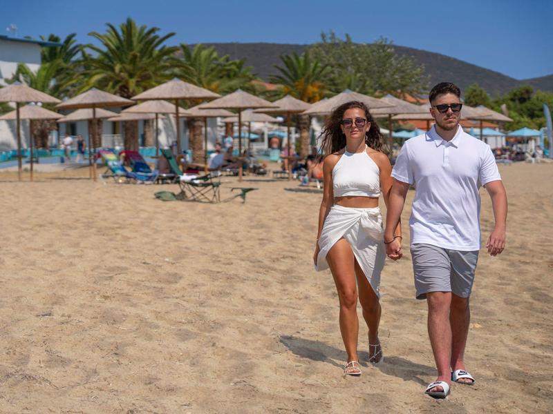 Para w letnich ubraniach spaceruje po piaszczystej plaży z parasolami i widokiem na basen.