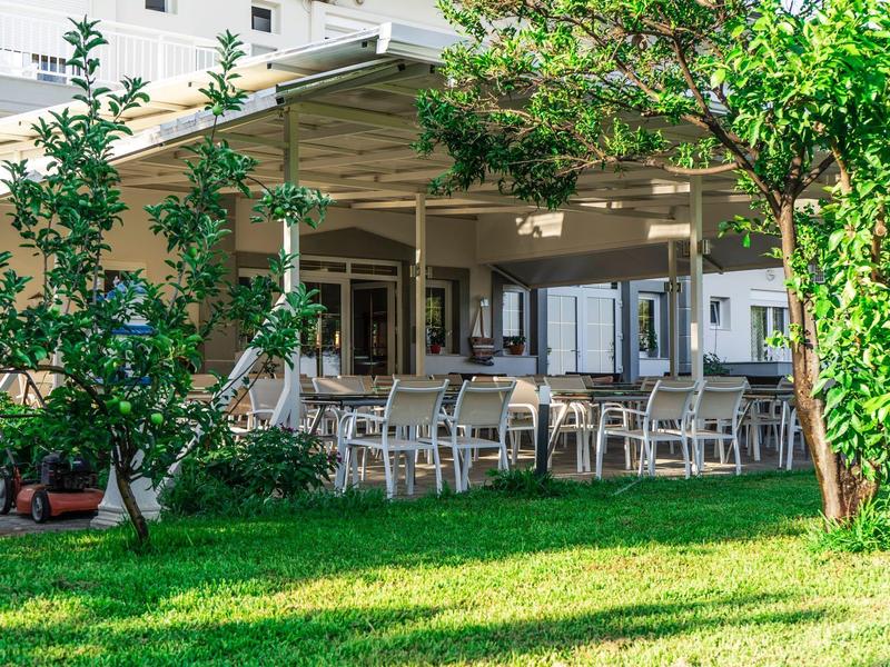 Taras hotelowy z krzesłami i stołami otoczony zieloną trawą i drzewami.