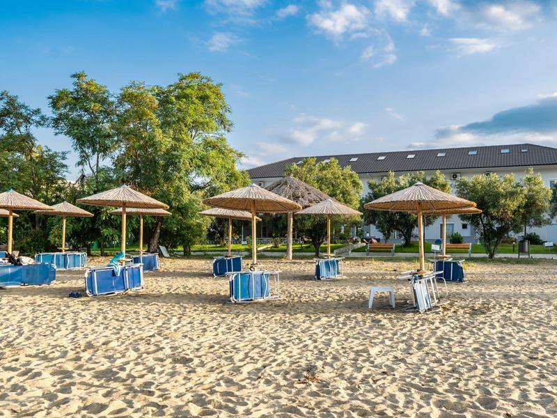 Plaża z piaskiem, niebieskimi leżakami i parasolami przed budynkiem hotelu pod niebieskim niebem.