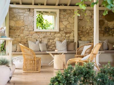 Przytulny teren zewnętrzny z kamienną ścianą, drewnianymi krzesłami i poduszkami pod zadaszonym tarasem.