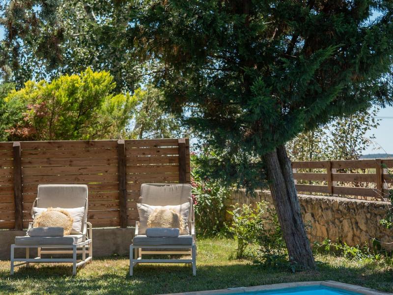 Two lounge chairs beside a pool under a large tree in a garden with a wooden fence.
