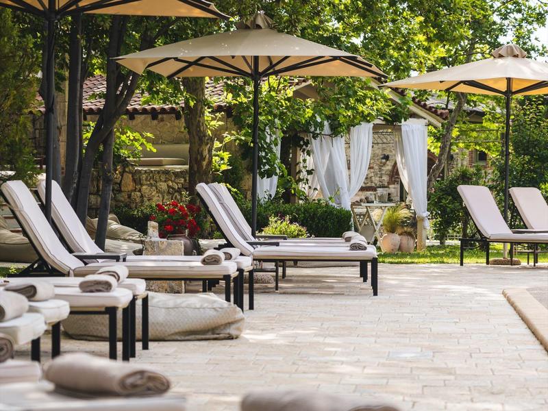 Several lounge chairs with towels under umbrellas by a pool in a green garden setting.