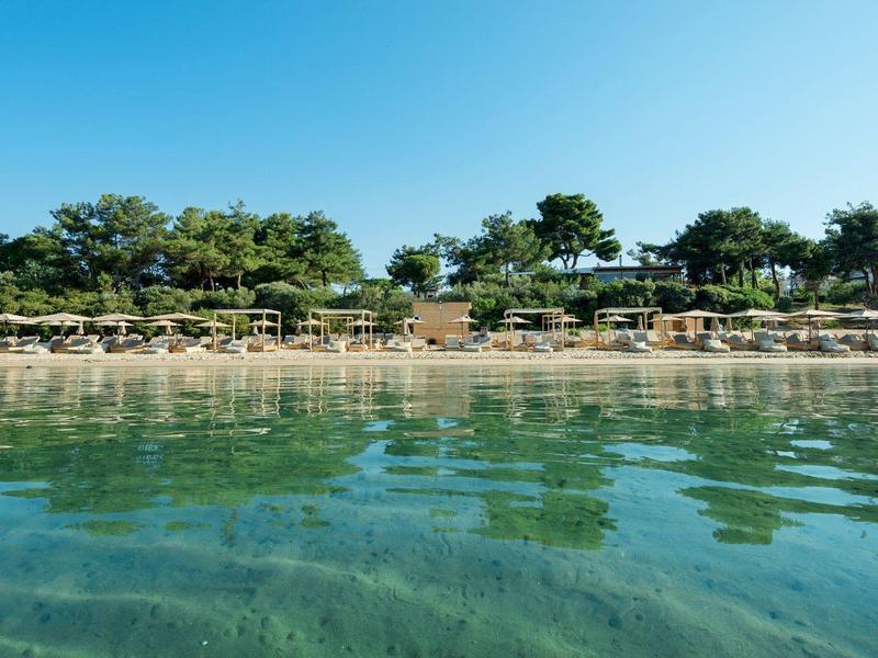 Spiaggia con lettini e ombrelloni, acqua limpida e cielo azzurro, circondata da vegetazione verde.